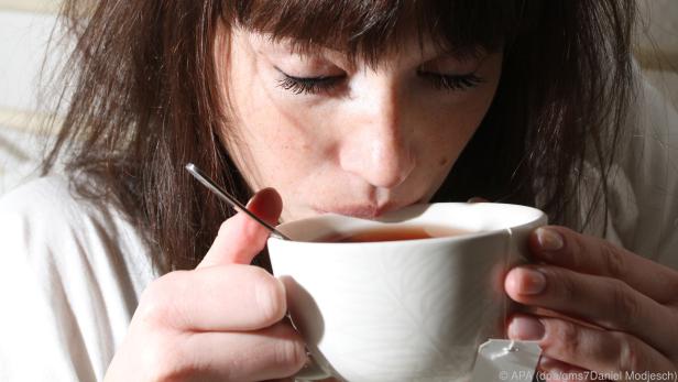 Ein Frau hält eine weiße Tasse Tee in Händen und setzt mit dem Mund zum ersten Schluck an.