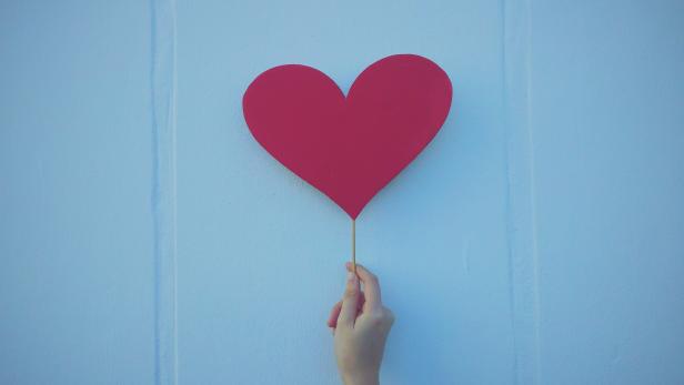 Eine rosa Herz auf einem Holzstiel wird von einer Hand an eine blaue Wand gehalten.