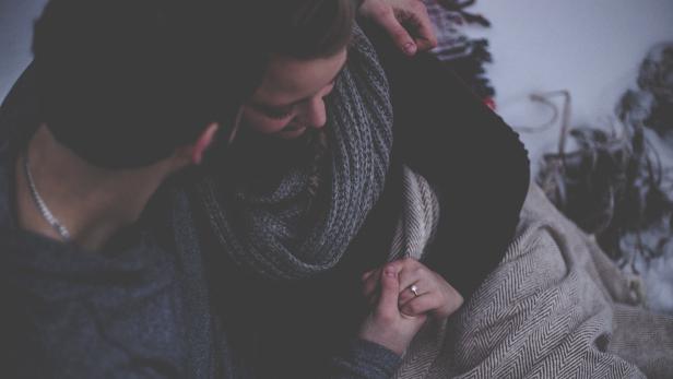 Ein Pärchen sitzt umarmt nebeneinander und halten Händchen. Sie sind mit einer Decke zugedeckt. 