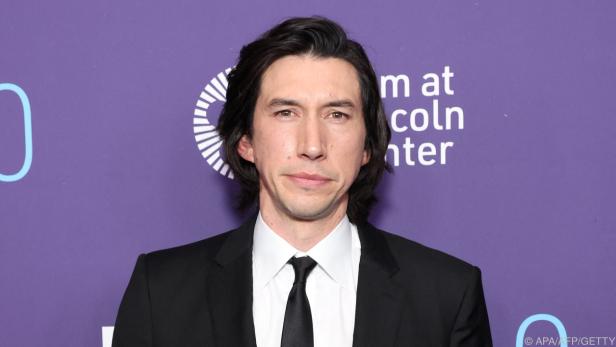 Adam Driver steht vor einer lila Werbewand. Der Schauspieler trägt einen schwarzen Anzug