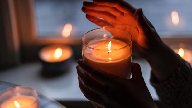 Person hält eine brennende Kerze in der Hand, mit der anderen Hand schützt sie die Flamme vor dem Ausgehen. Im Hintergrund sind weitere brennende Kerzen zu sehen. 