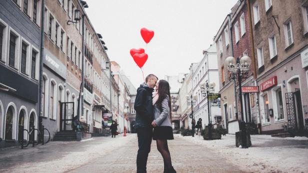 Ein Paar, das rote Herz-Luftballons hält, küsst sich leidenschaftlich inmitten einer Fußgängerzone