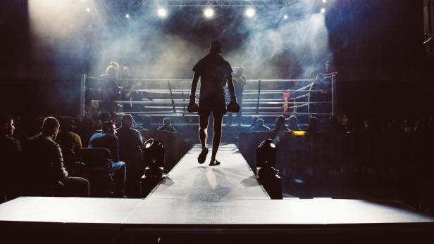 Ein Boxer auf dem Weg Richtung Boxring; links und rechts von ihm sitzt das Publikum