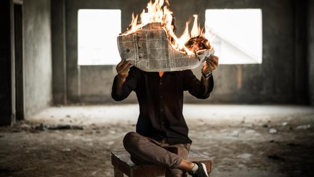 Mann sitzt in einem verlassenem Gebäude auf einem Stuhl. In seinen Händen hält er eine brennende Zeitung