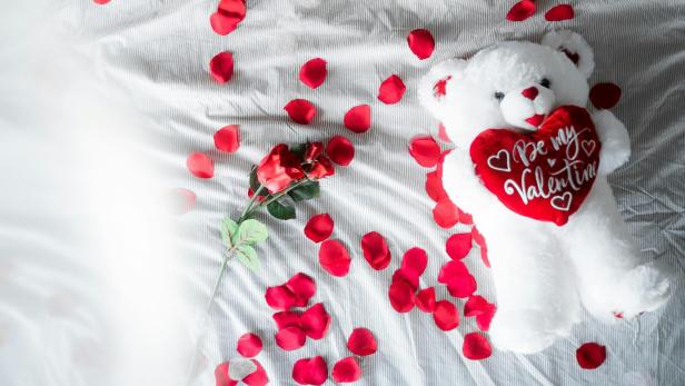 Weißer Teddy mit rotem Herz liegt zusammen mit Rosenblättern auf weißem Bettlaken
