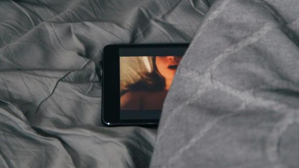 Tablet mit stöhnender Frau auf dem Bildschirm liegt inmitten grauer Bettlaken