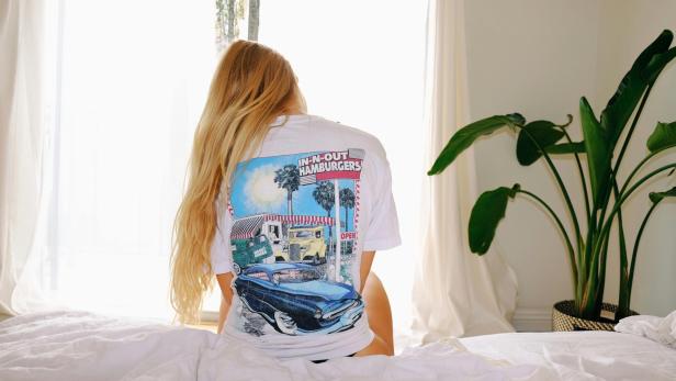 Blonde Frau sitzt mit dem Rücken zur Kamera auf einem Bett, sie trägt ein Shirt mit einem blauen Auto auf dem Rücken