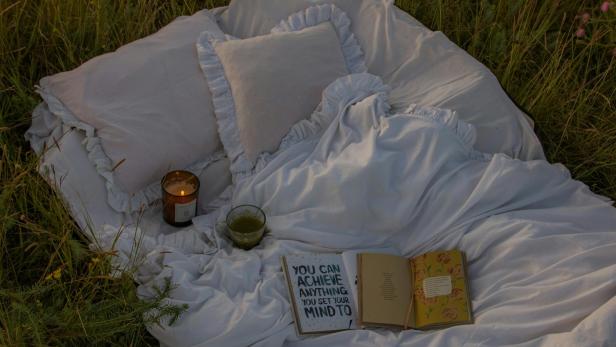 weiße Bettwäsche liegt zusammen mit einem Buch und einer Kerze auf einer Wiese