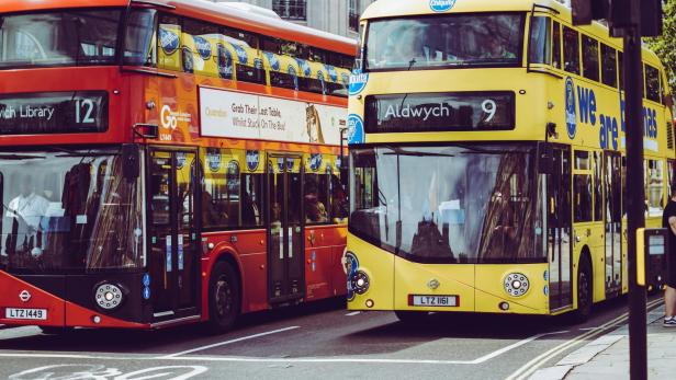 Ein roter Doppeldeckerbus steht links neben einem gelben an einer Ampel auf einer Straße in England.