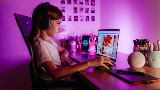 Langhaariges Mädchen sitzt mit Kopfhörern an einem Schreibtisch vor dem Laptop und schaut konzentriert