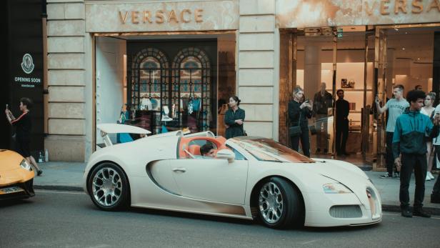 Ein creme-weißer Bugatti Veyron parkt vor einer Versace-Boutique.