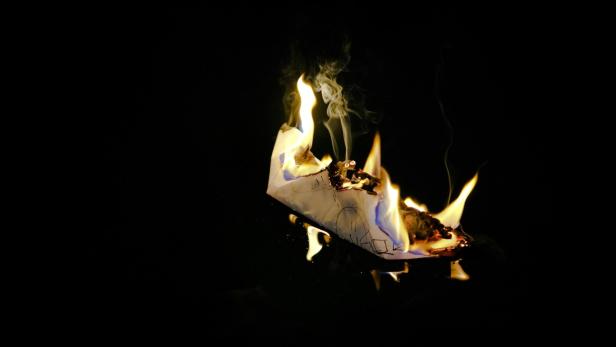 Ein gefaltetes Papier-Boot brennt vor einem schwarzen Hintergrund.
