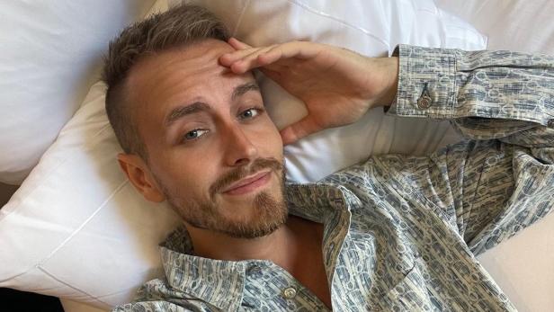 Blonder junger Mann liegt in gemustertem Hemd im Bett und macht ein Selfie